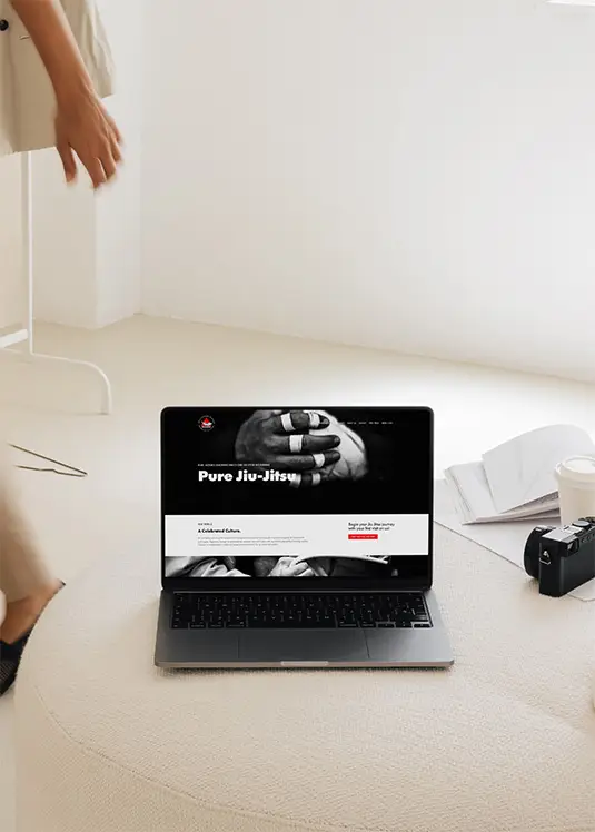 Laptop open on a clean ottoman, showing a bold Jiu-Jitsu website