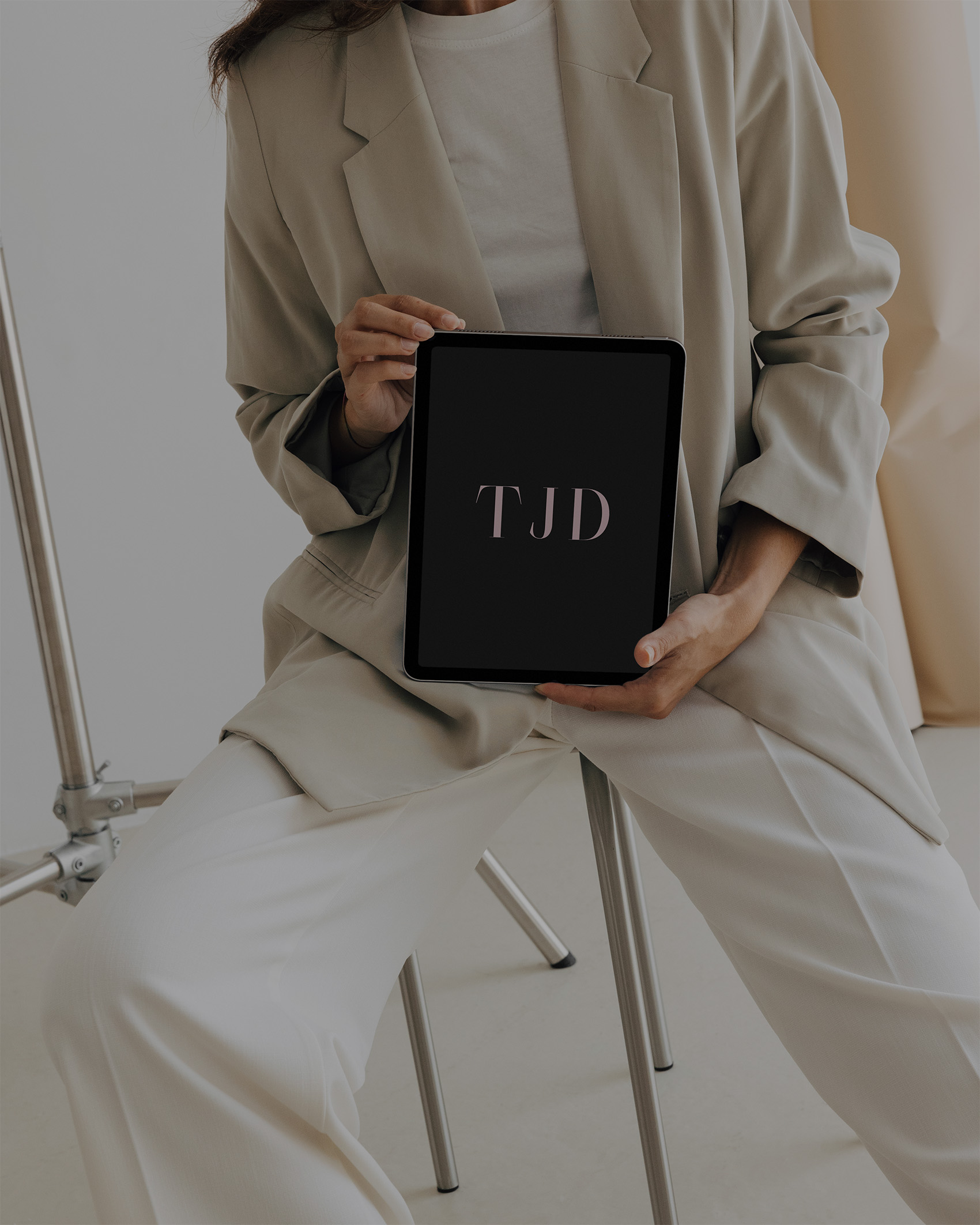 Professional portrait of Tina Jenner, founder of TJD design studio, holding iPad displaying TJD branding, seated in minimalist neutral setting