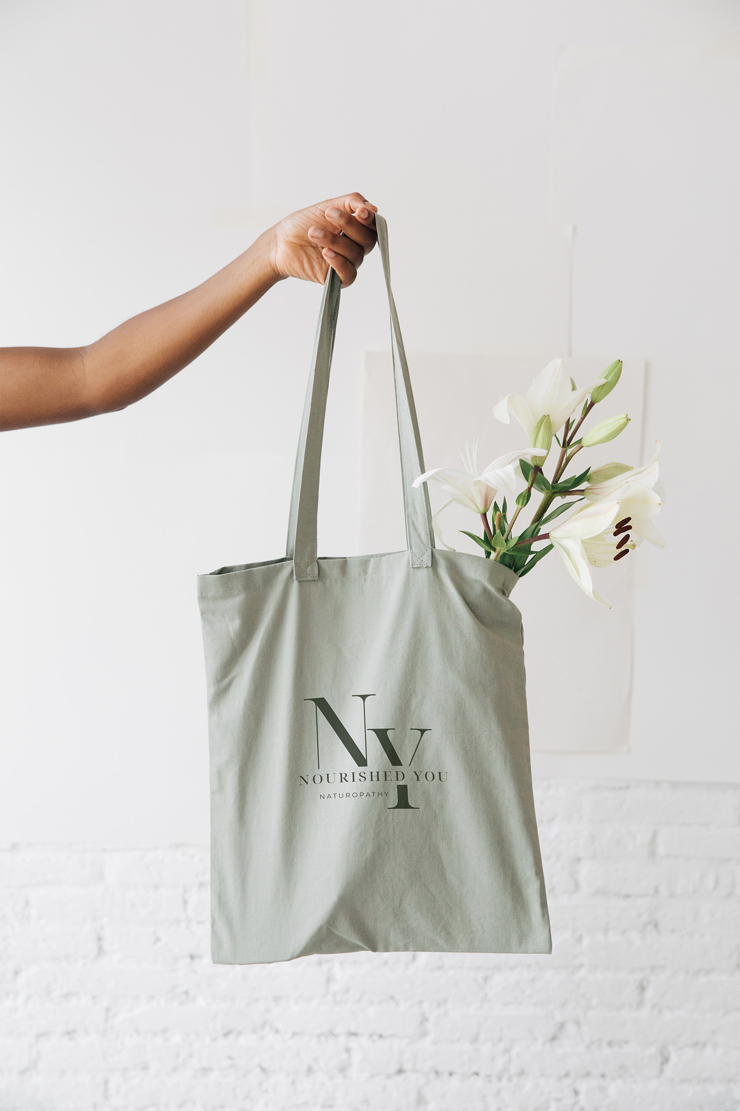 Nourished You branded sage green tote bag with logo, held with white flowers, brand extension merchandise for naturopath practice
